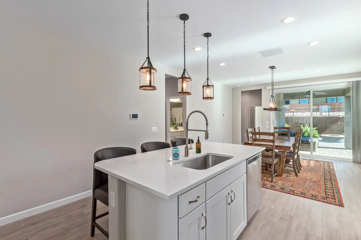 A warm, communal kitchen scene with an island sink, bar stools, hanging light fixtures, and a dining table in the background. The space is designed for cooking, entertaining, and family gatherings, exuding a sense of togetherness.