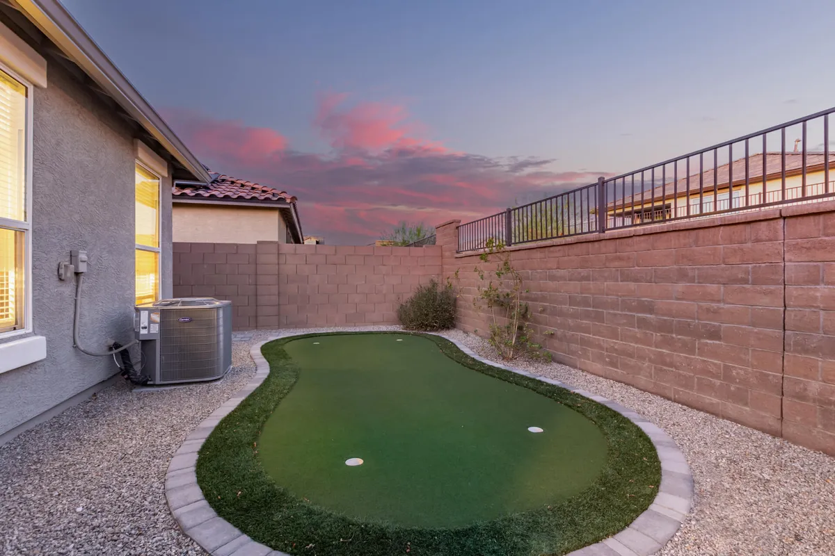A tranquil exterior scene featuring an artificial putting green surrounded by decorative gravel and landscaped walls. The setting is bathed in soft sunset light, creating a peaceful ambiance ideal for relaxing or practicing golf. The area is designed for small gatherings and personal enjoyment.