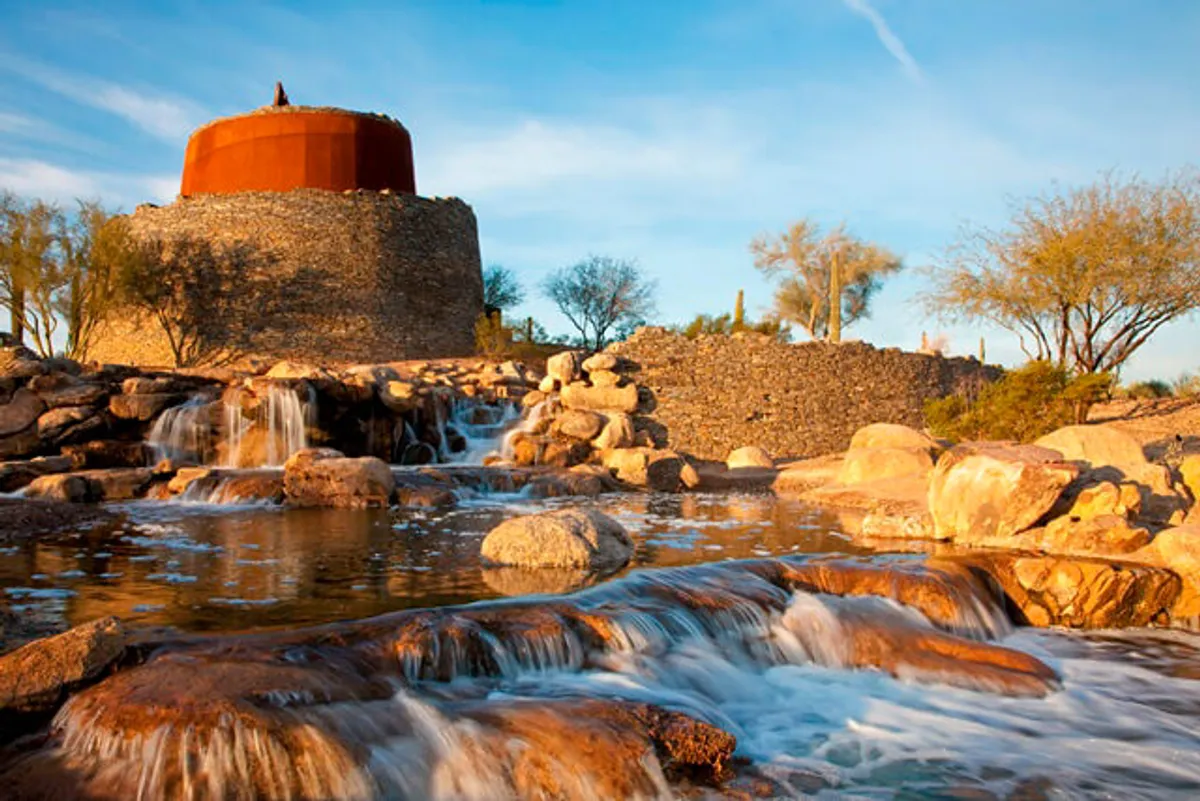 A serene landscape with a stone structure set beside flowing water, surrounded by lush vegetation and dramatic rock formations. The scene offers opportunities for nature walks, relaxing by the water, and exploring scenic desert trails, with golden light suggesting a sunset or early morning adventure.