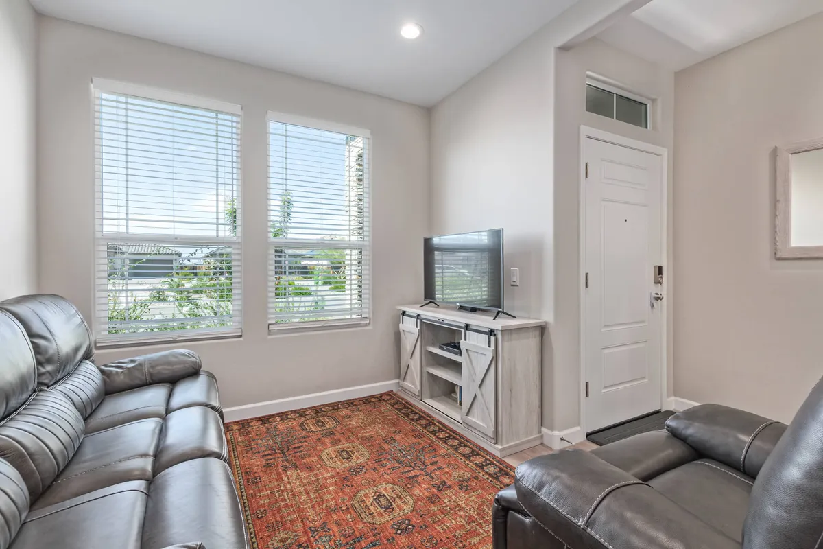 This interior image features a comfortable living room with a leather sofa facing a TV stand and television, complemented by an area rug. Natural light streams in through windows with blinds, creating a bright and inviting space. The arrangement encourages relaxation and socializing, with cozy movie nights and game day gatherings as natural uses.