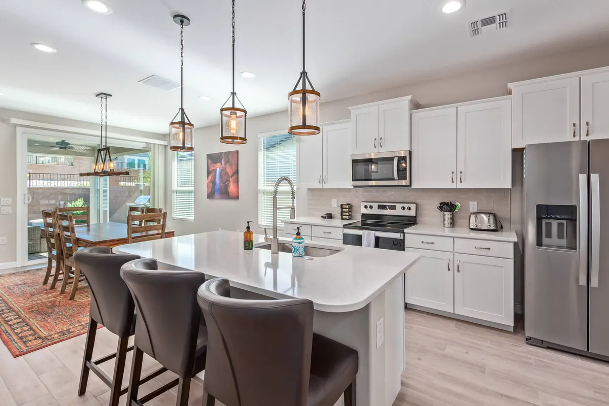This image captures an open-concept interior where a stylish island countertop with hanging pendant lights leads seamlessly to a dining area framed by large windows. The space is well-lit and designed for entertaining, cooking, and casual dining, embodying a welcoming and contemporary mood.