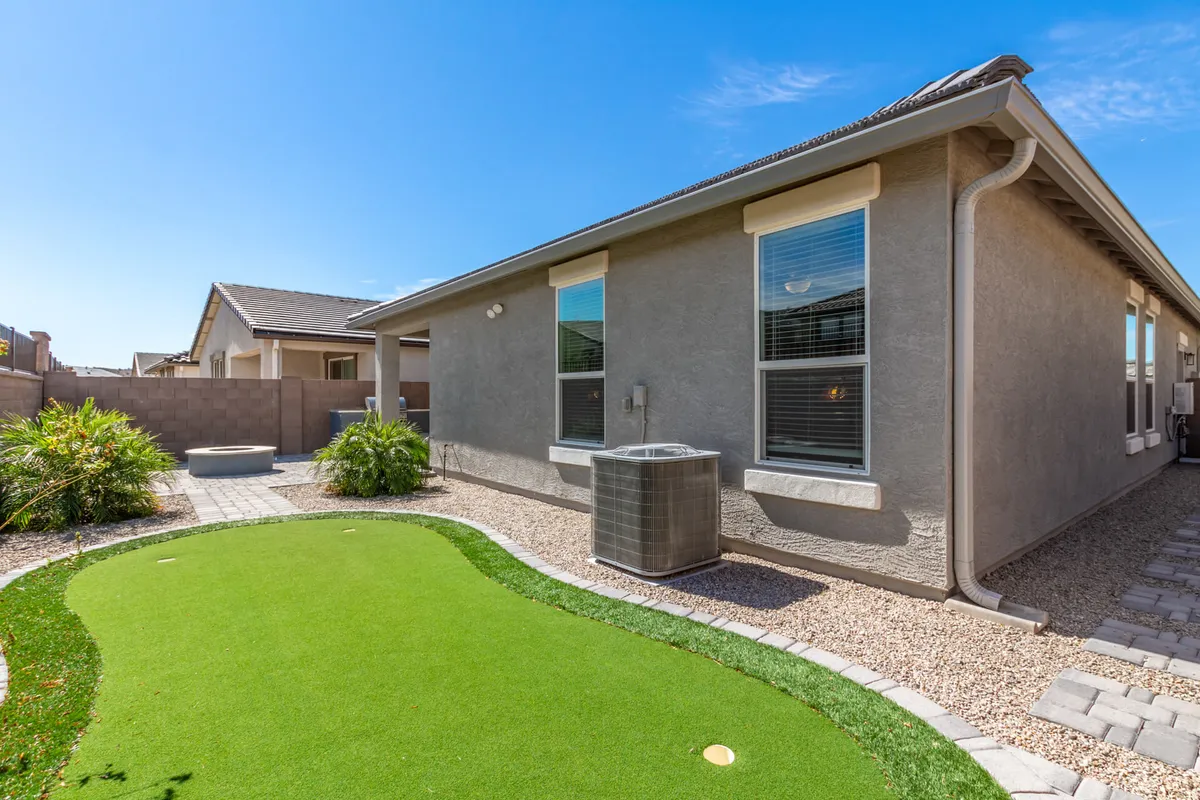 A beautifully landscaped backyard with an artificial putting green, lush garden elements, and the home’s exterior wall in view. The space invites playful outdoor activities like golf, relaxed gatherings, and quiet afternoons in the sun, highlighted by vibrant greenery and comfortable lounging spots.