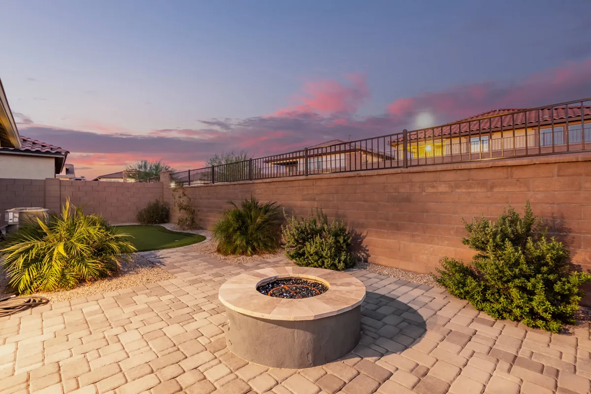 A cozy paved seating area surrounds a glowing fire pit, framed by lush greenery and a privacy fence. The setting is serene, encouraging intimate conversations or moments of relaxation under the stars, with palm trees and inviting landscaping setting the Arizona ambiance.