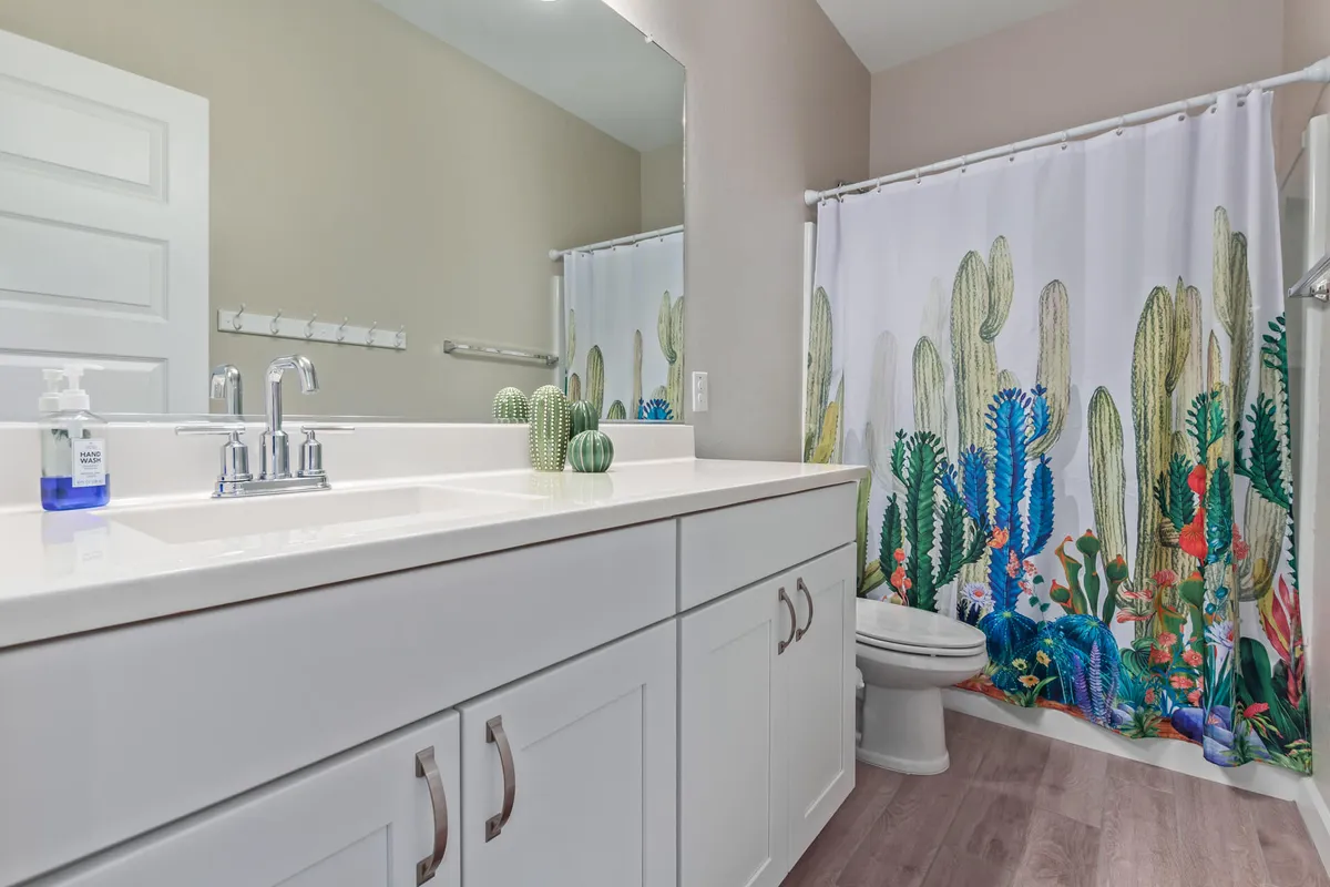 A bathroom with a modern vanity, dual sinks, and a bright, colorful shower curtain featuring a cactus design. Clean decor and cheerful lighting make the space feel fresh and inviting.