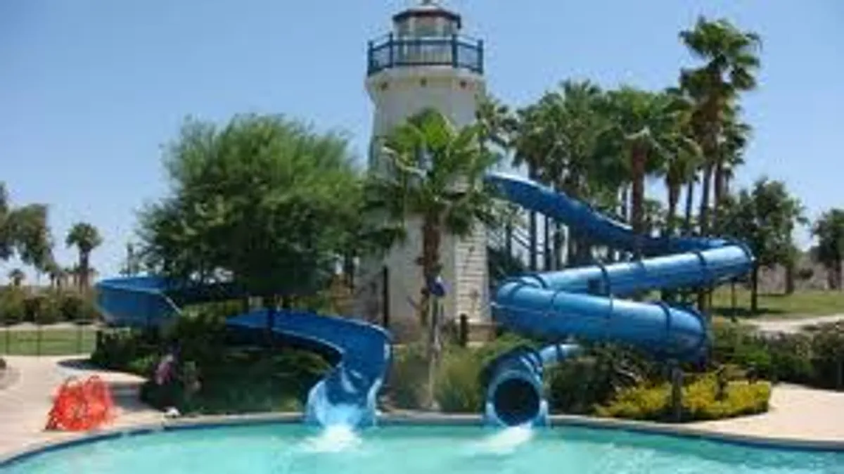 The image showcases the home's private backyard oasis, featuring a shimmering swimming pool, twisting water slides, a whimsical lighthouse structure, and tall, lush palm trees. Sunlight sparkles on the water as families and friends enjoy swimming, sliding, lounging under the palms, and embracing the outdoor fun.