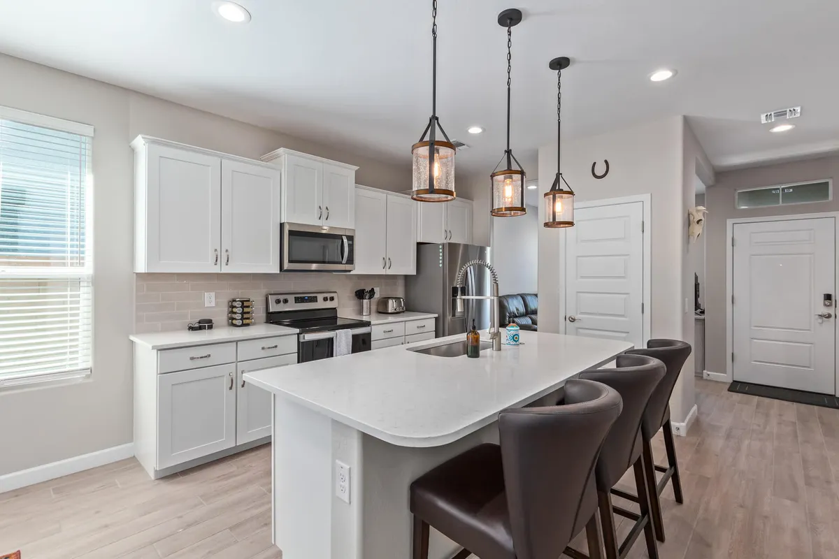 This image features a modern kitchen with an island countertop, pendant lights, stainless steel appliances, bar stools, and large windows that fill the space with natural light. The setting feels open and welcoming, ideal for both cooking and casual dining.