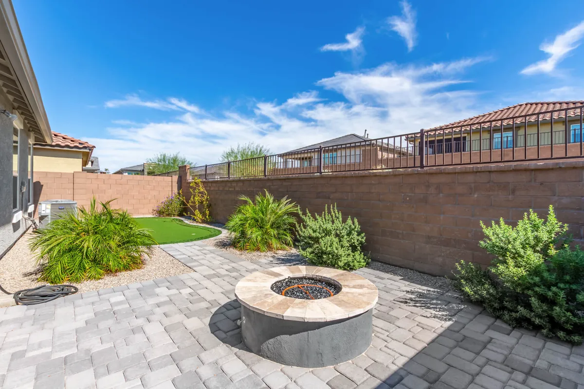 An inviting outdoor space features a fire pit surrounded by landscaped greenery and paver stones. The setup is ideal for both stargazing in the evening and savoring coffee in the morning, with various seating options that encourage year-round enjoyment.